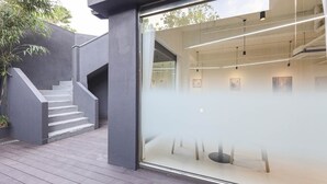 Ein modernes Gebäude mit einer Treppe, die zu einer Glastür führt, einer Holzterrasse und Blick ins Innere, wo Deckenleuchten und Kunstwerke zu sehen sind.