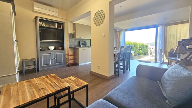 Una sala de estar con un sofá, una mesa de centro de madera y un comedor con sillas y una mesa. Se observa una cocina con armarios y un refrigerador.