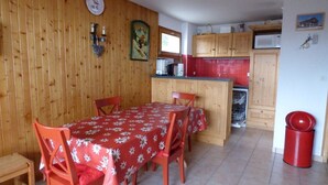 Un coin repas avec une table et des chaises en bois, une cuisine avec des placards en bois et une poubelle rouge.