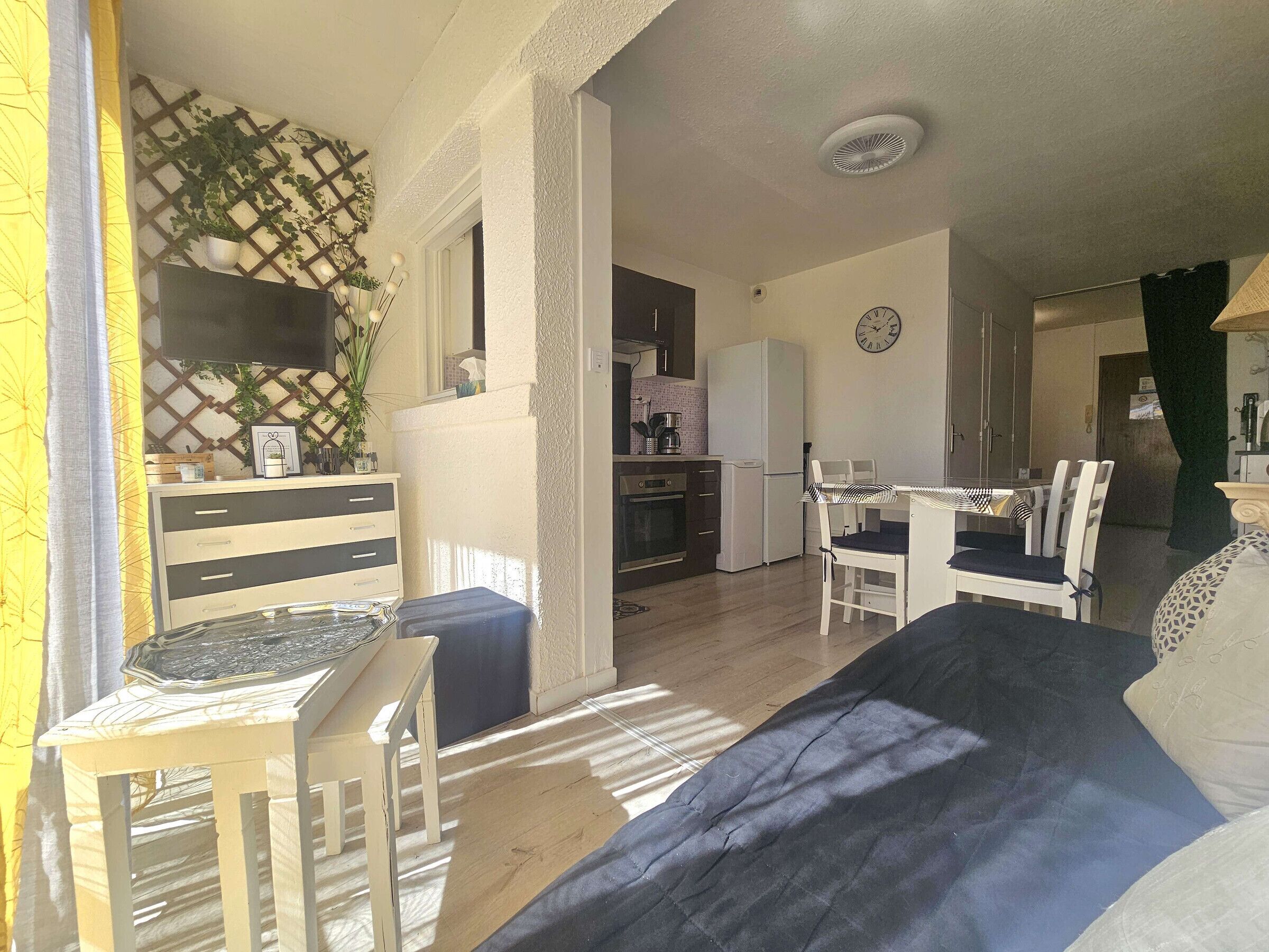 A cozy room with a bed, a small table, and chairs. There is a kitchen area with a refrigerator and a clock on the wall.