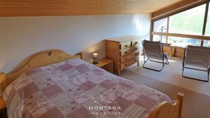 A bedroom with a wooden ceiling, a bed with a floral bedspread, a nightstand with a lamp, a wooden dresser, two chairs, and a window with a view of the outdoors.