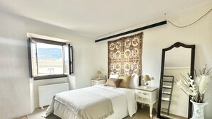 A bedroom with a large bed, a nightstand, a mirror, and a window with a view of buildings. - Medieval House in the Center of Montblanc (Montblanc)