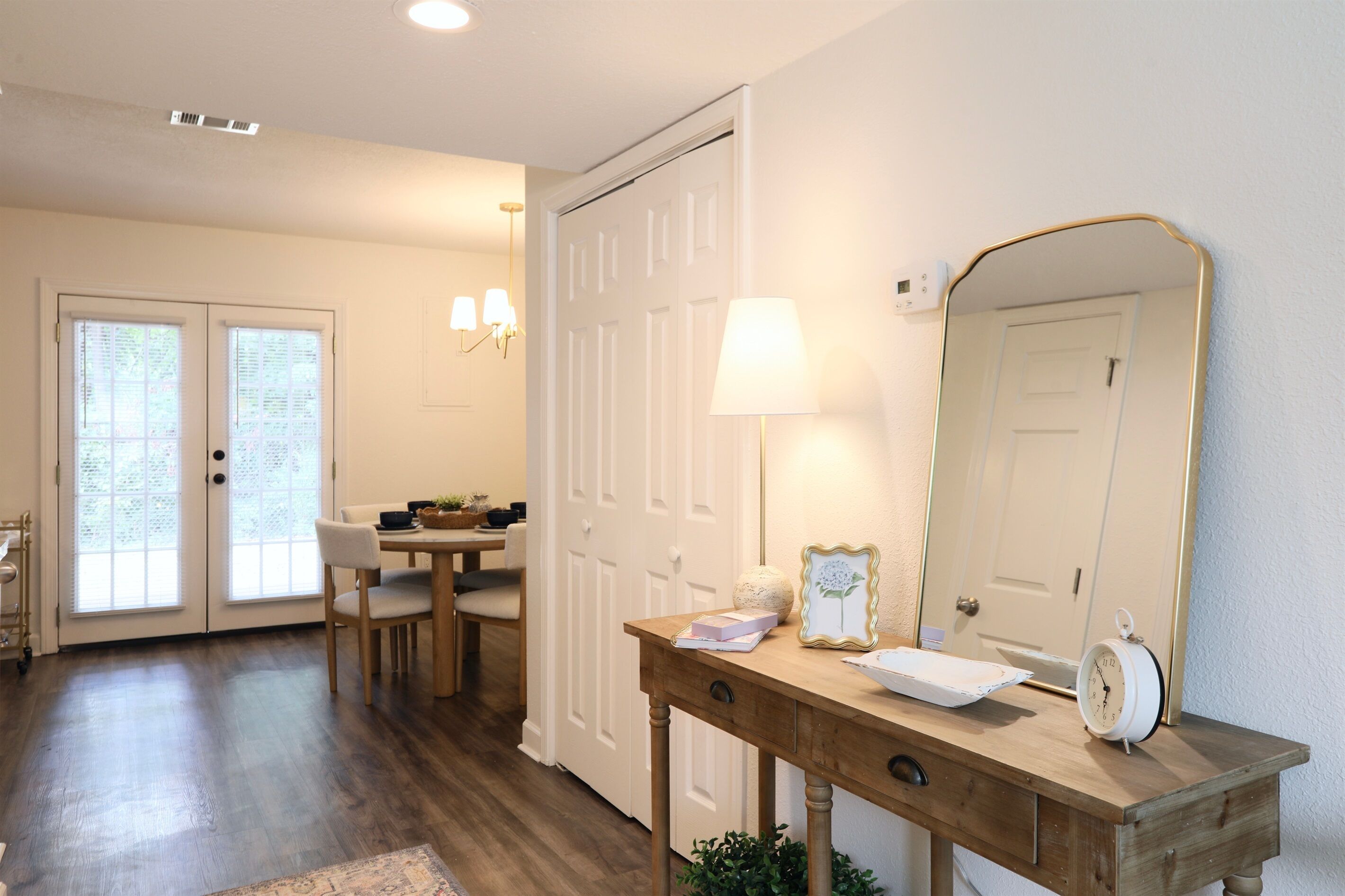 Una habitación con un escritorio de madera, un espejo, una lámpara y un reloj. Se ve una puerta con un ventanal y una silla.