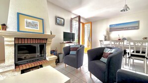 Un salon avec une cheminée, deux fauteuils, un téléviseur et un coin repas comprenant une table et des chaises.