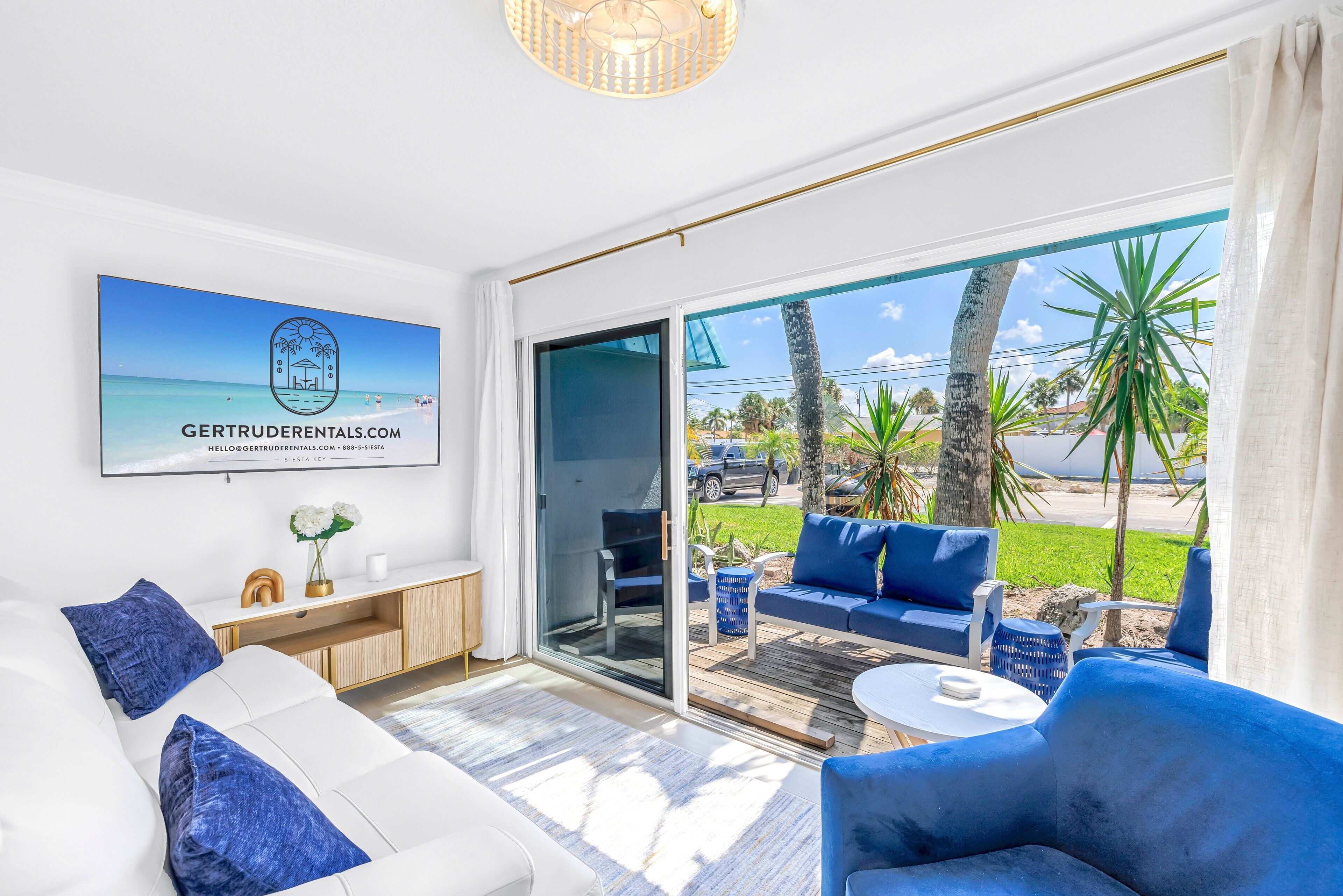 A modern living room with a sofa, blue armchairs, and a coffee table. There is a large window with a view of palm trees and a beach. A framed picture of a beach scene is on the wall.