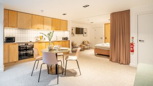 Une cuisine moderne avec des meubles en bois, un coin repas avec une table ronde et des chaises, une chambre avec un lit et des rideaux, et un extincteur visible.