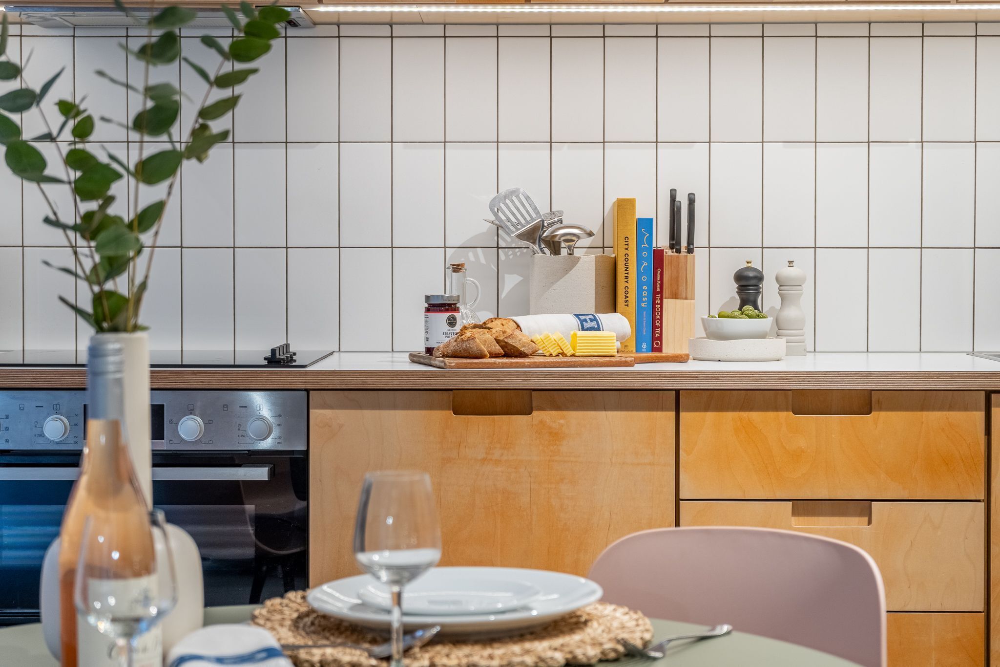 Une cuisine avec un dosseret de carreaux blancs, des armoires en bois, un plan de travail agrémenté d’un panier à pain, d’un verre à vin et d’un vase avec une plante.