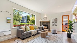 A modern living room with a grey sofa, a fireplace, a large window with a view of buildings and trees, and a wooden floor. - Modern 2BR in DT Renton, 3 AC, WS 95, Shop&Parks (Renton)