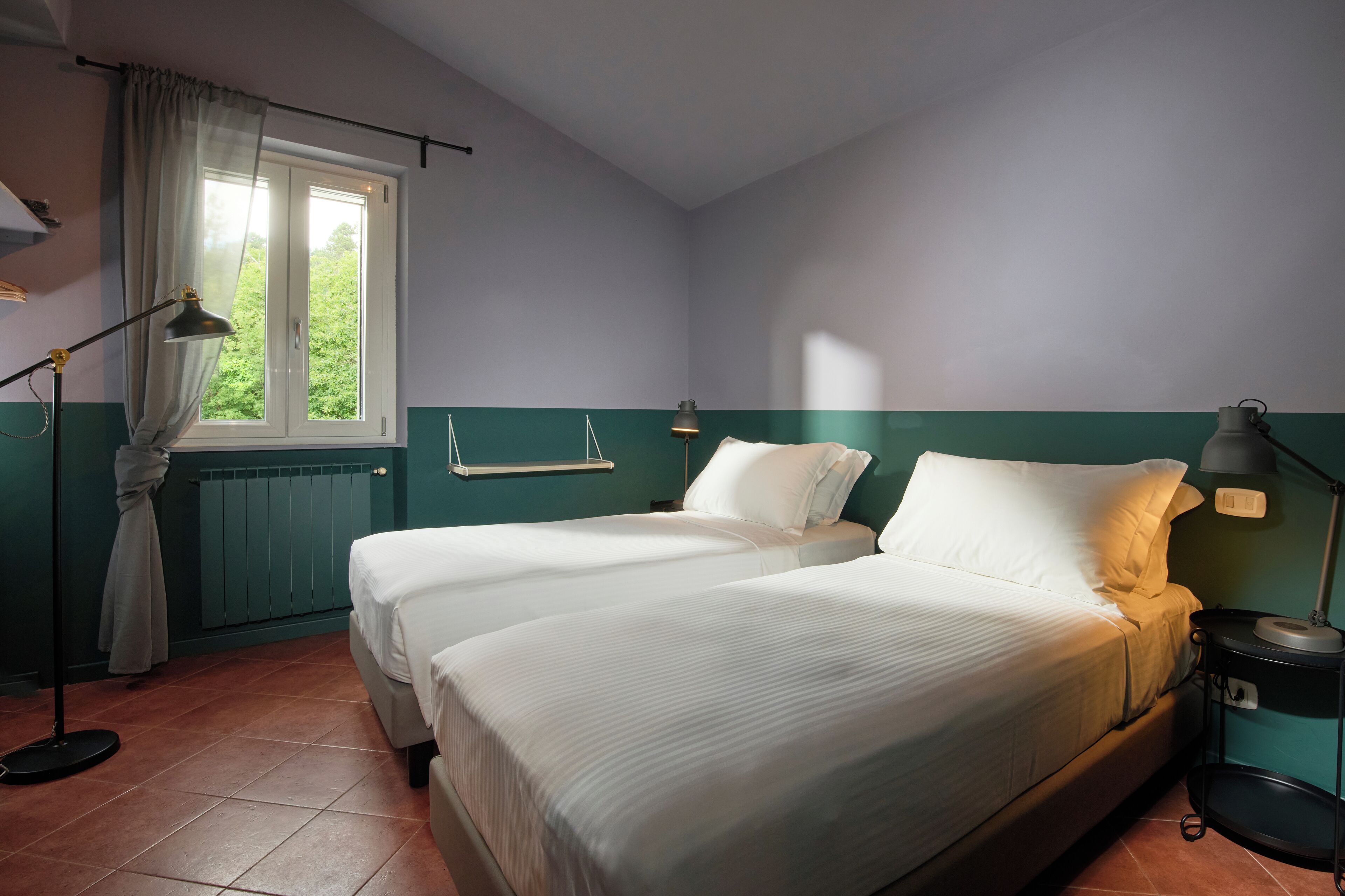 A room with two beds, a window with curtains, a radiator, and a nightstand with a lamp.