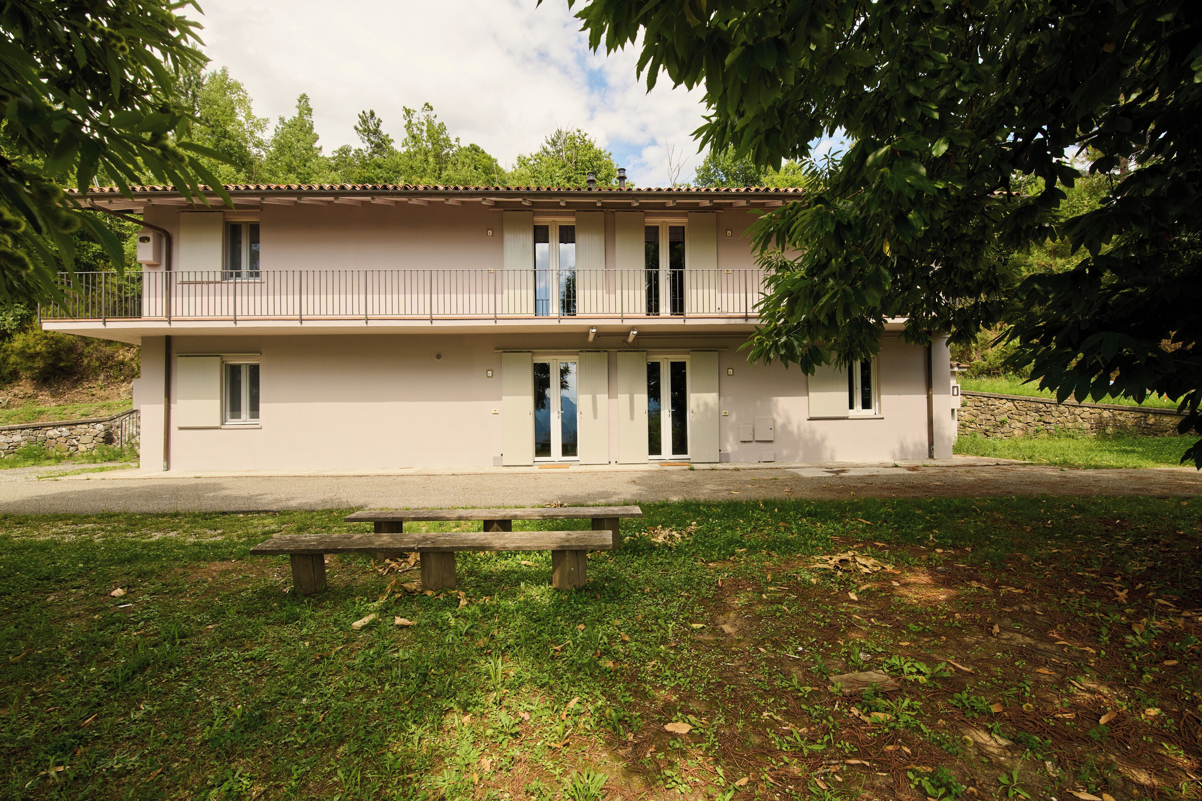 A two-story building with a balcony, a bench in front, and a stone wall in the background.