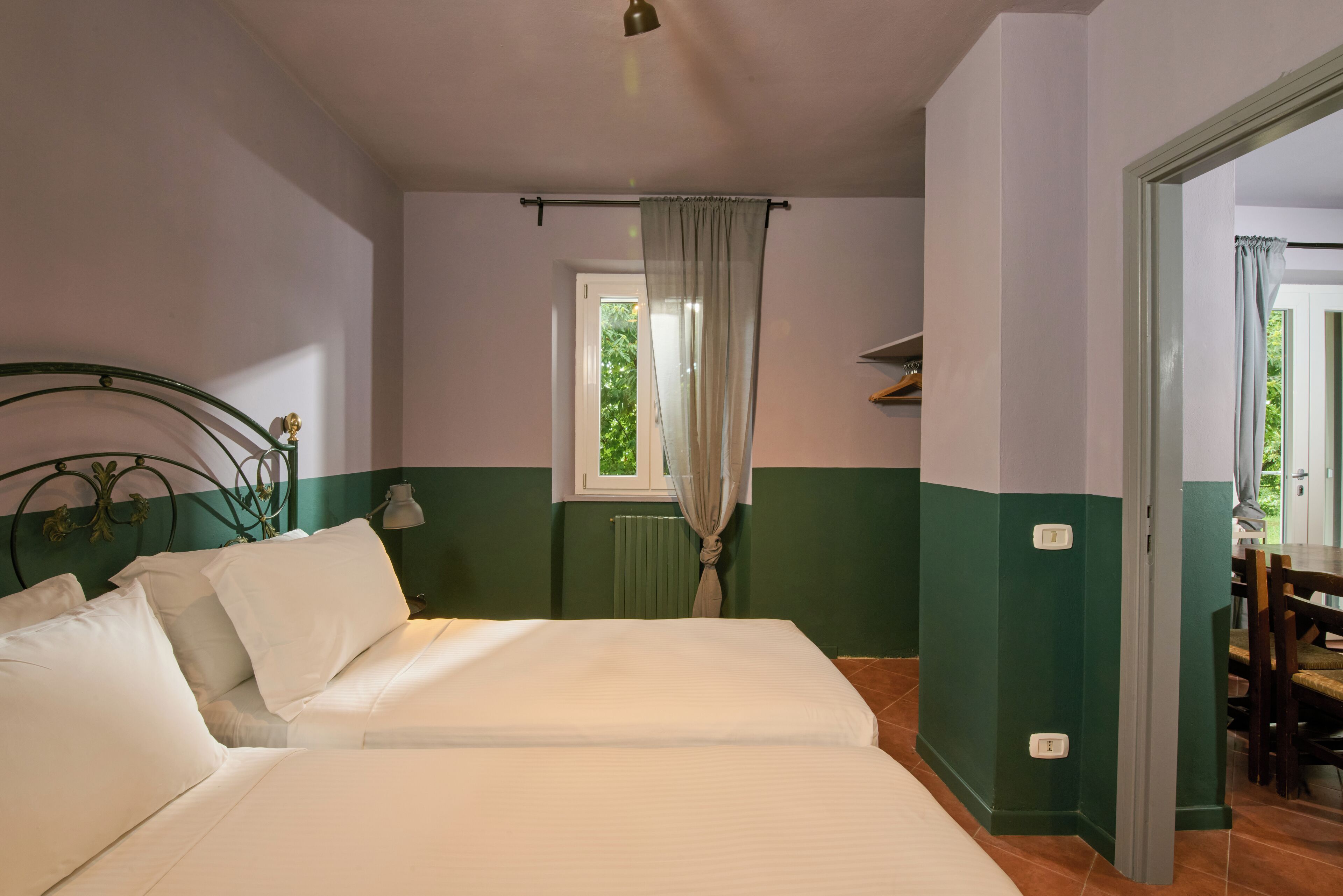 A bedroom with two beds, a window with curtains, a radiator, and a door leading to another room.