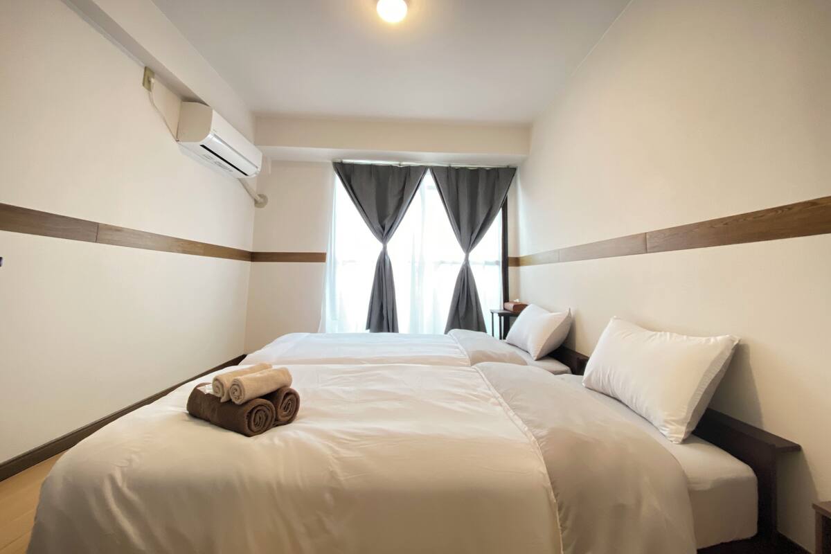 A hotel room with two beds, a ceiling light, an air conditioning unit, and a window with curtains.