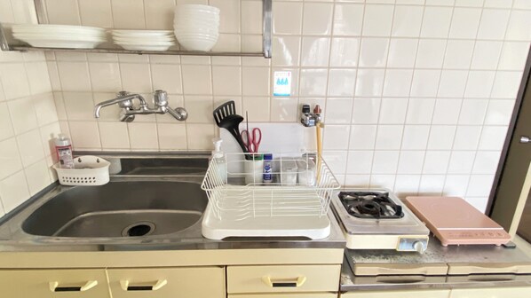A kitchen with a sink, stove, and various utensils. There are plates and bowls on a shelf above the sink. - AkiraHome Tsutenkaku (Osaka)
