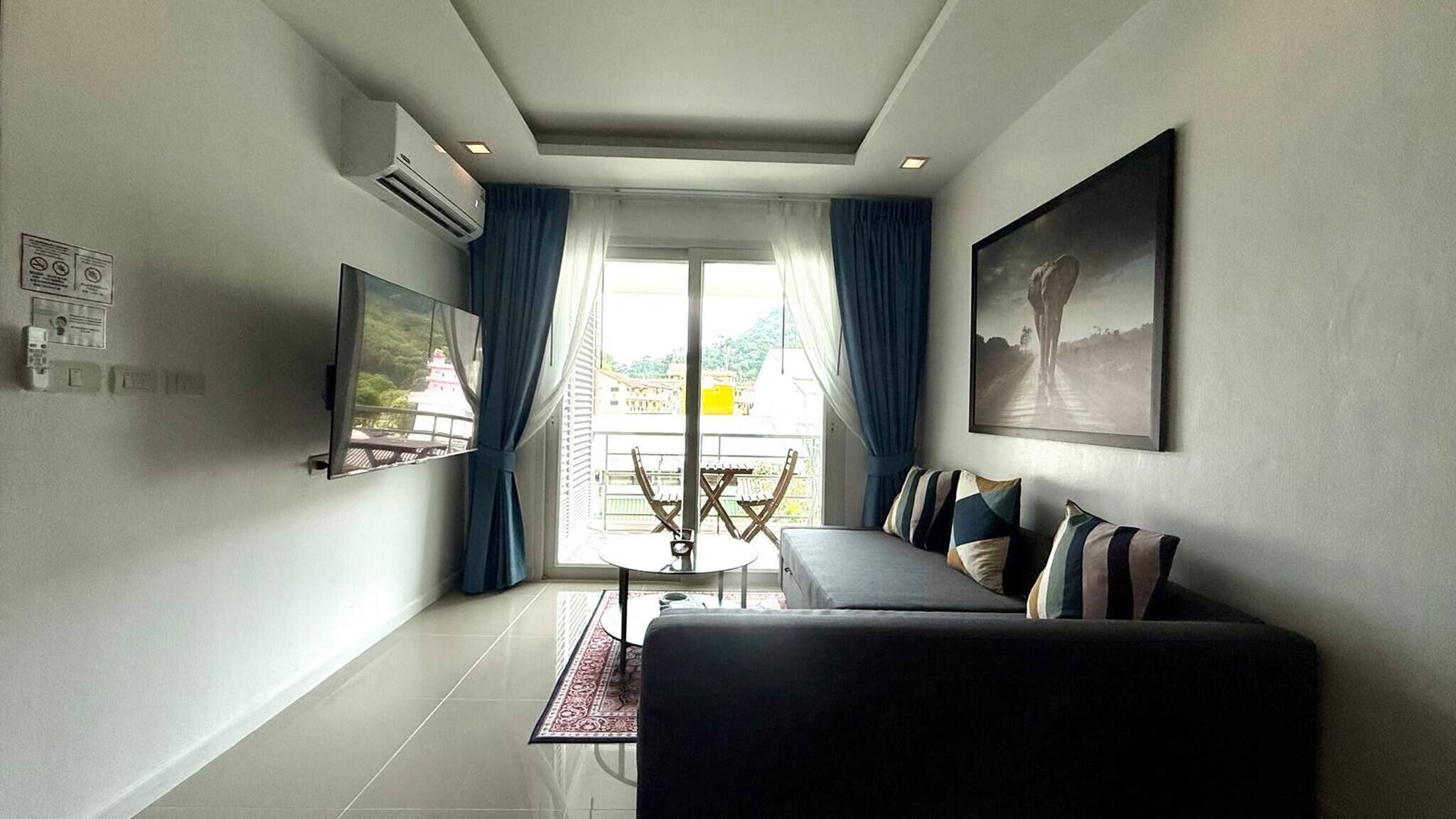 A modern living room with a sofa, a dining table, and a TV. There is a balcony with a view of buildings and a tree.