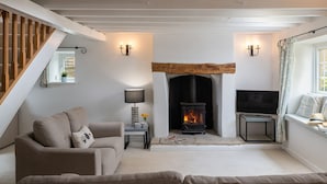 A cozy living room with a sofa, a small table with a lamp, a fireplace, and a TV. - Murray's Cottage - sleeps 5 guests  in 3 bedrooms (Bourton-on-the-Water)