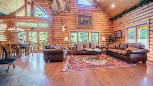 A log cabin interior with a wooden floor, leather sofas, a dining area, and a large window.