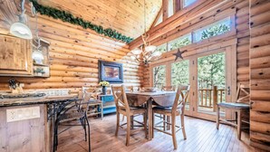 A rustic wooden cabin interior with a dining area, kitchen, and large windows offering a view of the outdoors.
