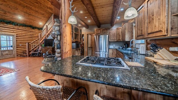A rustic cabin interior with a stone pillar, wooden staircase, kitchen area with granite countertop, and a dining area with wicker chairs.