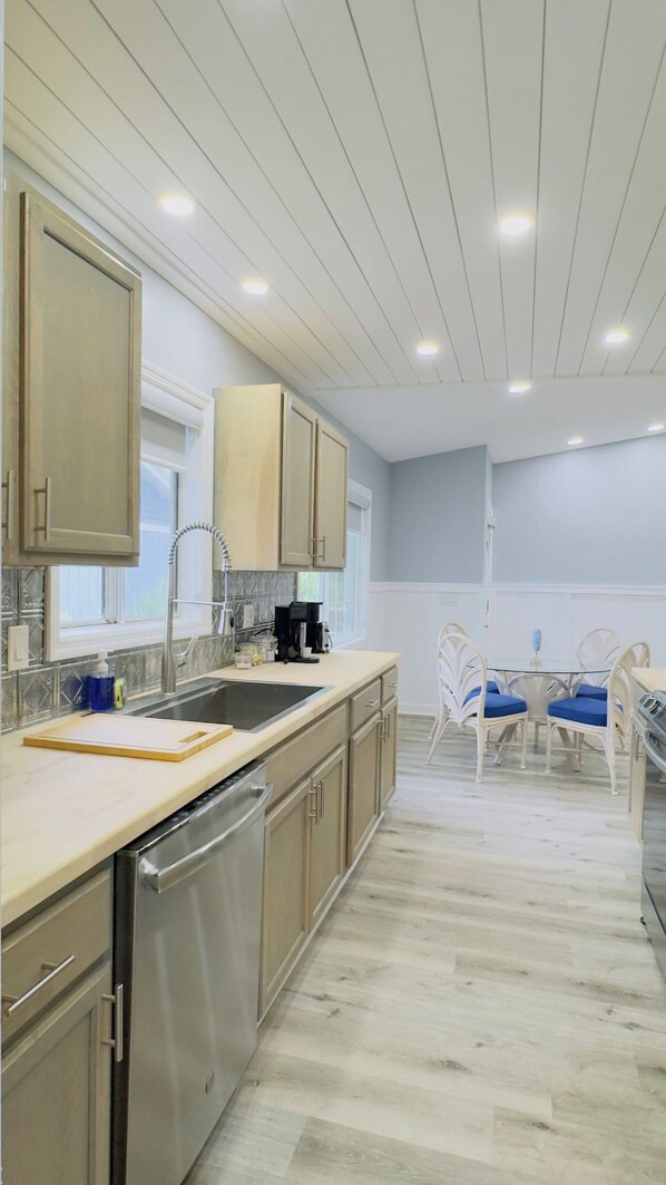 A modern kitchen with a dishwasher, stainless steel appliances, and a dining area with a glass table and white chairs. - Bright & airy 2BR, 2BA beach retreat A (North Myrtle Beach)