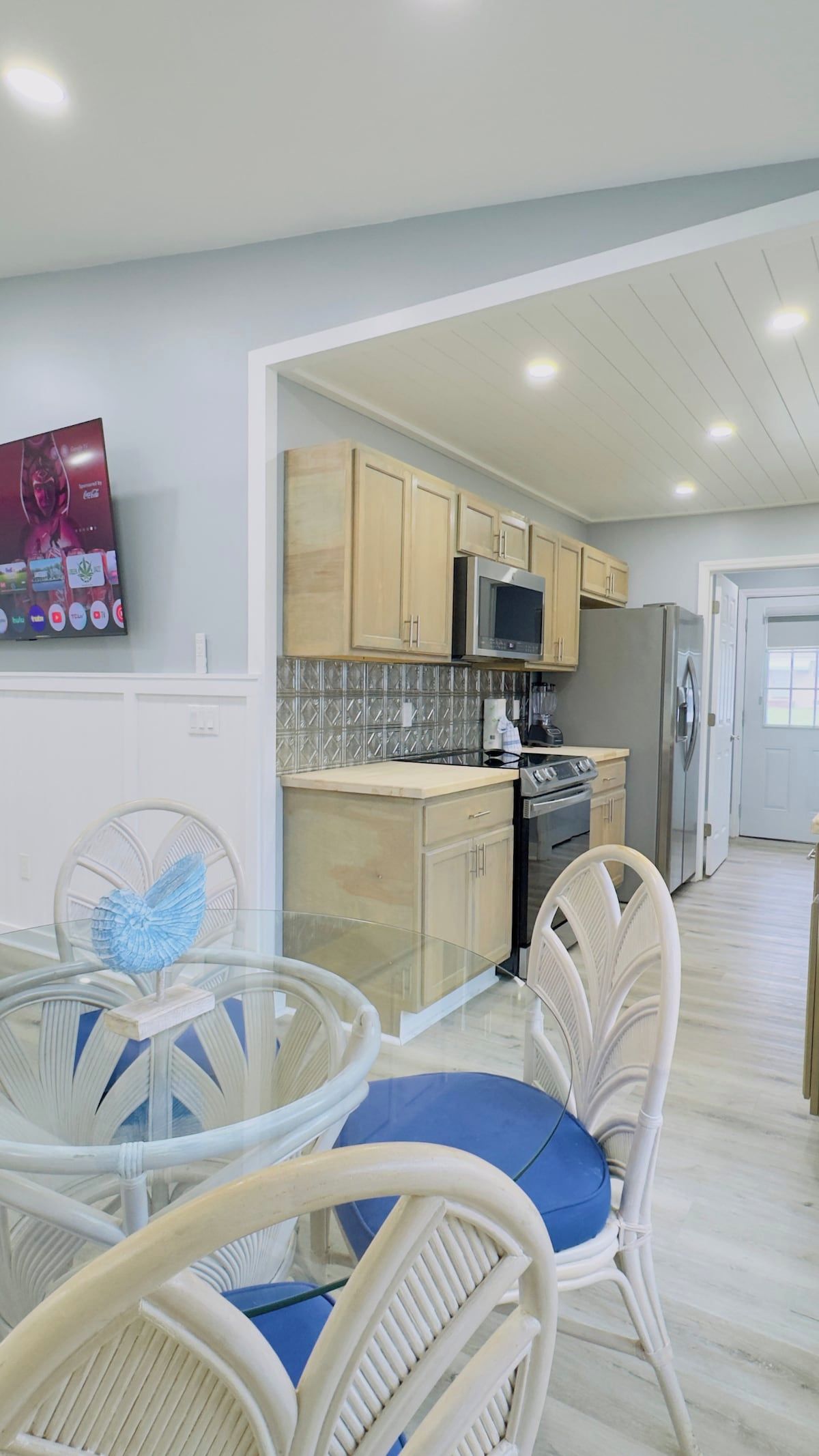 Una cocina moderna con armarios de madera, un refrigerador de acero inoxidable y un microondas. Hay una mesa de vidrio con sillas y un objeto decorativo sobre la mesa.