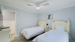 Une chambre avec deux lits, un ventilateur de plafond, une table de chevet avec une lampe et un tableau au mur.