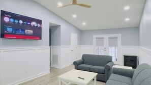 Una sala de estar con un televisor de pantalla plana montado en la pared, un sofá gris, una mesa de centro blanca y una chimenea.