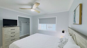 Une chambre à coucher comprenant un lit, une commode, un téléviseur, un ventilateur de plafond, une fenêtre avec des stores et un tableau accroché au mur.