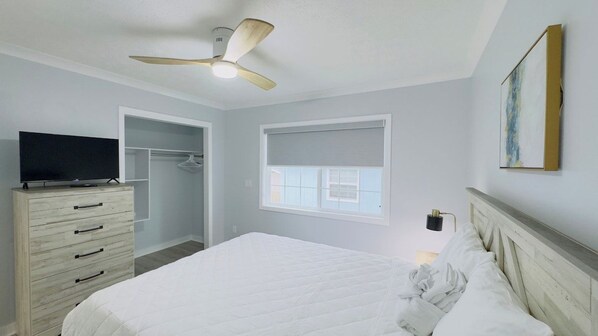 A bedroom with a bed, a dresser, a television, a ceiling fan, a window with blinds, and a painting on the wall.