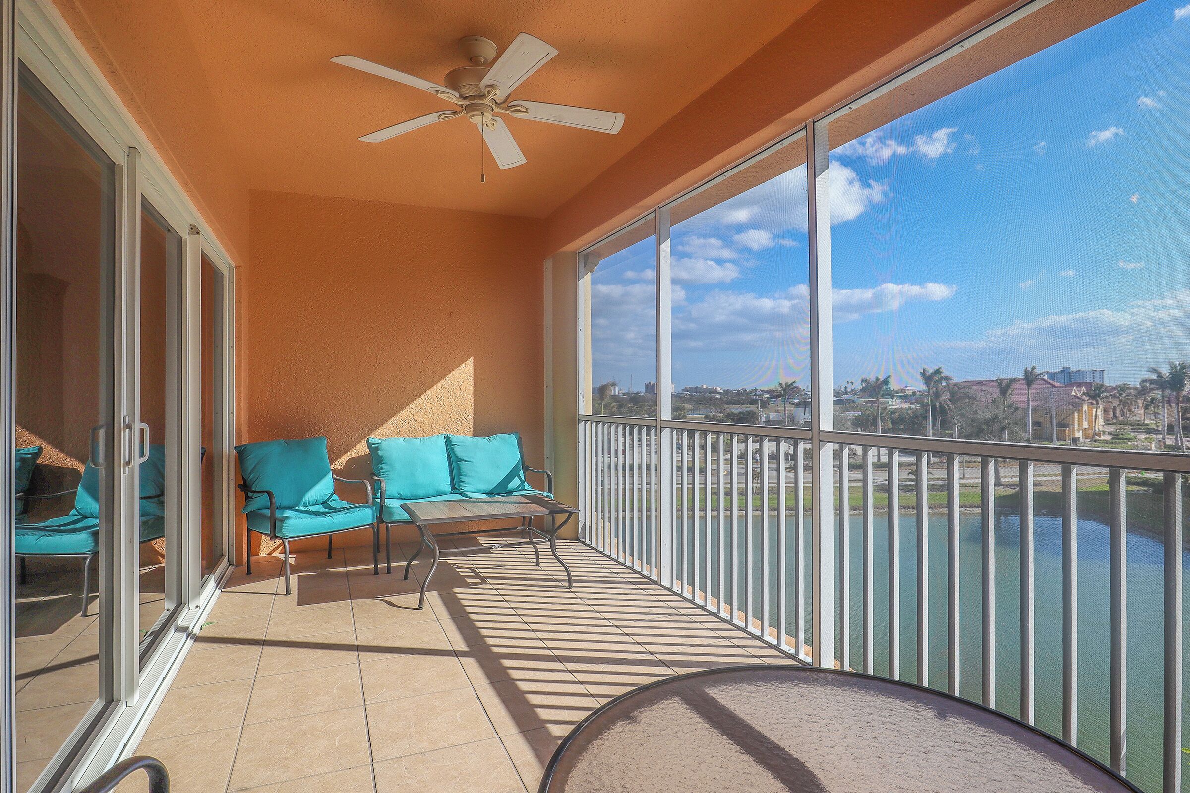 A balcony with a ceiling fan, a seating area with chairs and cushions, and a view of a body of water and buildings.