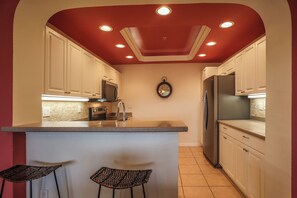 Una cocina con armarios blancos, una isla central con dos taburetes de bar, un refrigerador de acero inoxidable y un reloj de pared.