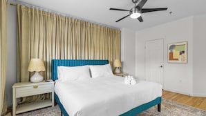 Una cama bien tendida con un cabecero azul, dos lámparas de noche, un ventilador de techo y una alfombra estampada.