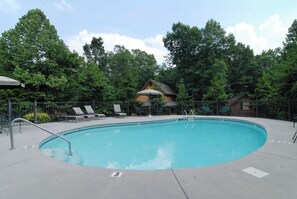 A round swimming pool with a metal ladder, surrounded by a fence. There are lounge chairs and a small building with a roof in the background.