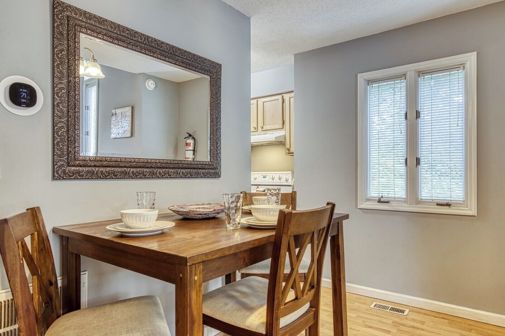 Un comedor con una mesa de madera, cuatro sillas, un espejo grande y decorativo, y una ventana con persianas.
