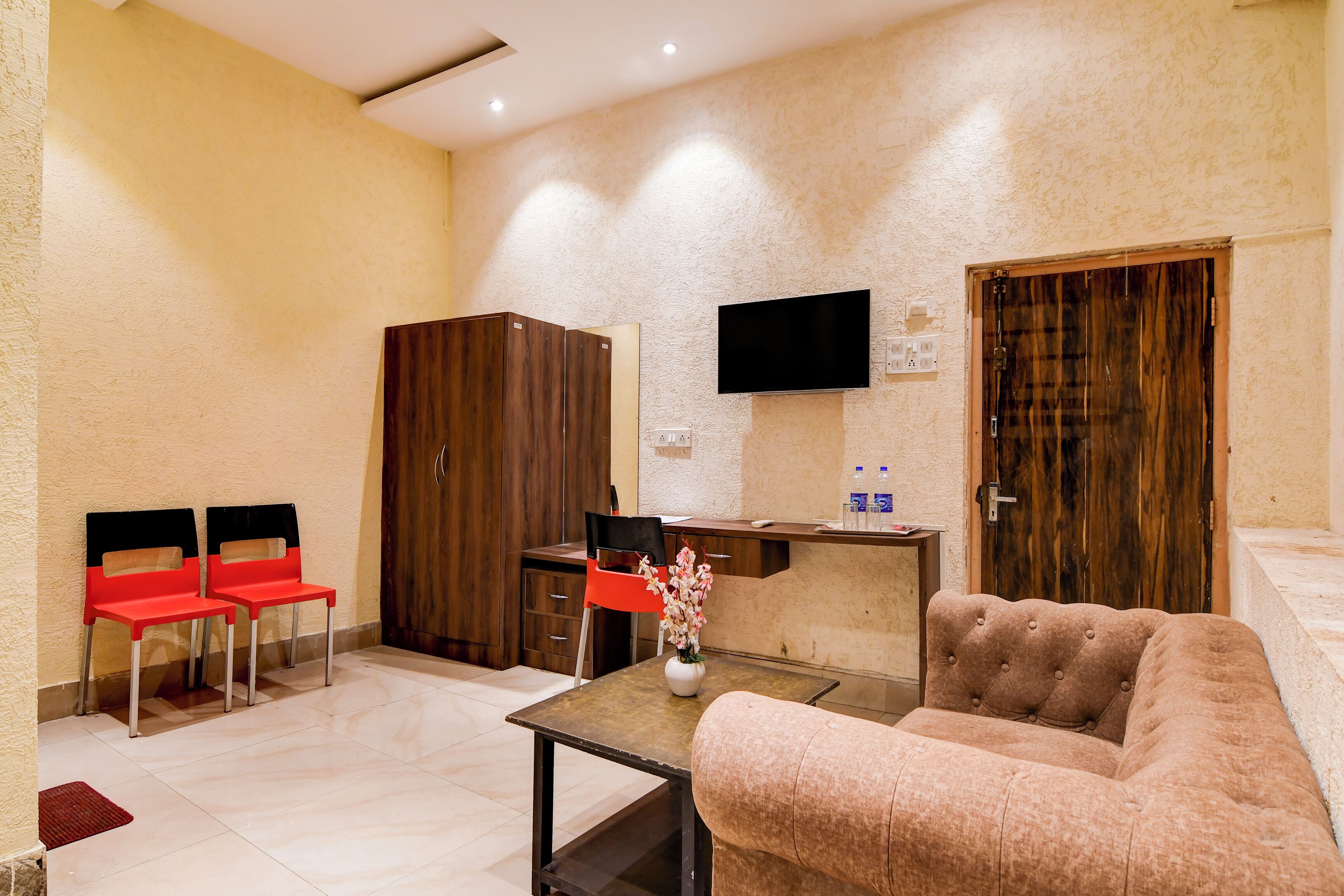 A hotel room with a sofa, a table with a vase, two red chairs, a wooden wardrobe, a desk with a chair, a TV mounted on the wall, and a wooden door.