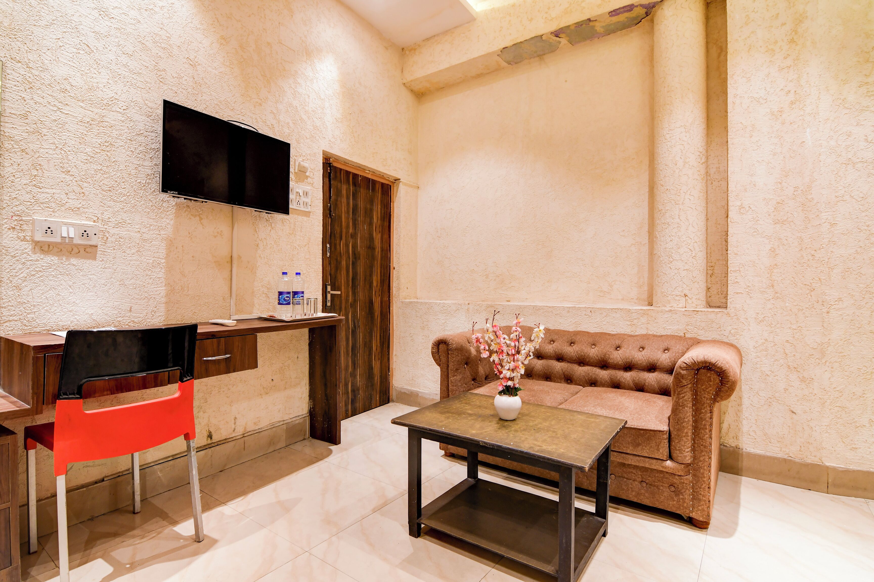 Una habitación con un sofá marrón, una silla roja, un televisor montado en la pared, una mesita con un florero y una puerta de madera.