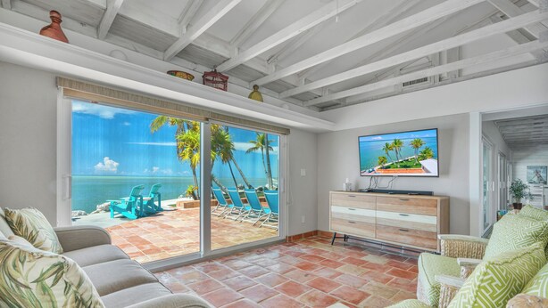 A living room with a large sliding glass door leading to a patio with a view of the ocean. There is a television on a wooden cabinet, and the room has a modern design with white walls and a tiled floor. - *NEW!*Paradise Point~VIEWS!~Open Water~Dock~Pool! (Marathon)