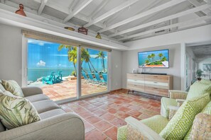 A living room with a large sliding glass door leading to a patio with a view of the ocean. There is a television on a wooden cabinet, and the room has a modern design with white walls and a tiled floor. - *NEW!*Paradise Point~VIEWS!~Open Water~Dock~Pool! (Marathon)