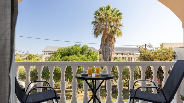 Ein Balkon mit zwei schwarzen Stühlen, einem kleinen runden Tisch sowie zwei Tassen Kaffee und Orangensaft. Im Hintergrund sind eine Palme und Gebäude zu sehen.