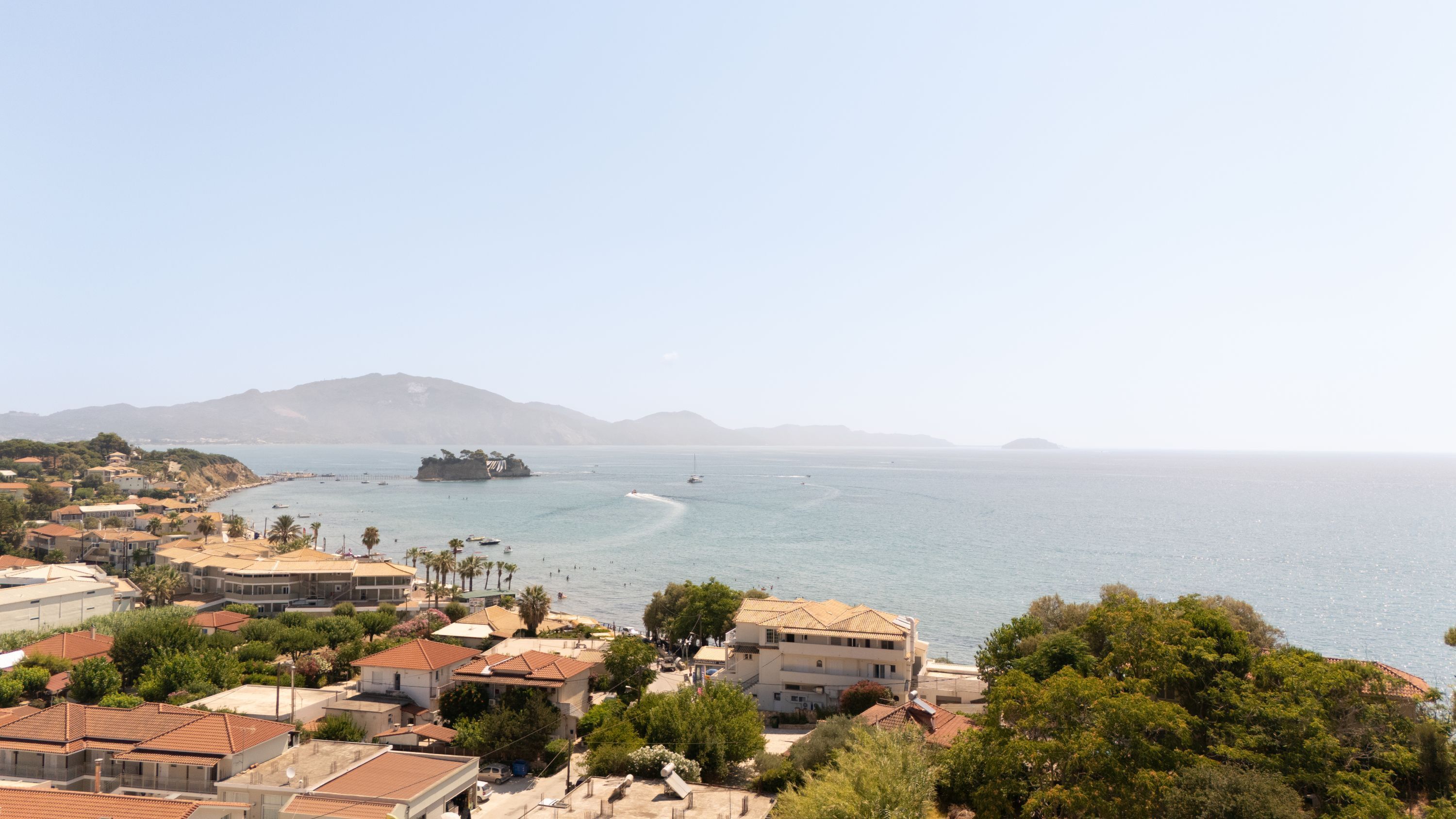 A coastal town with Mediterranean-style buildings, a clear view of the sea, and a prominent rocky island in the distance.