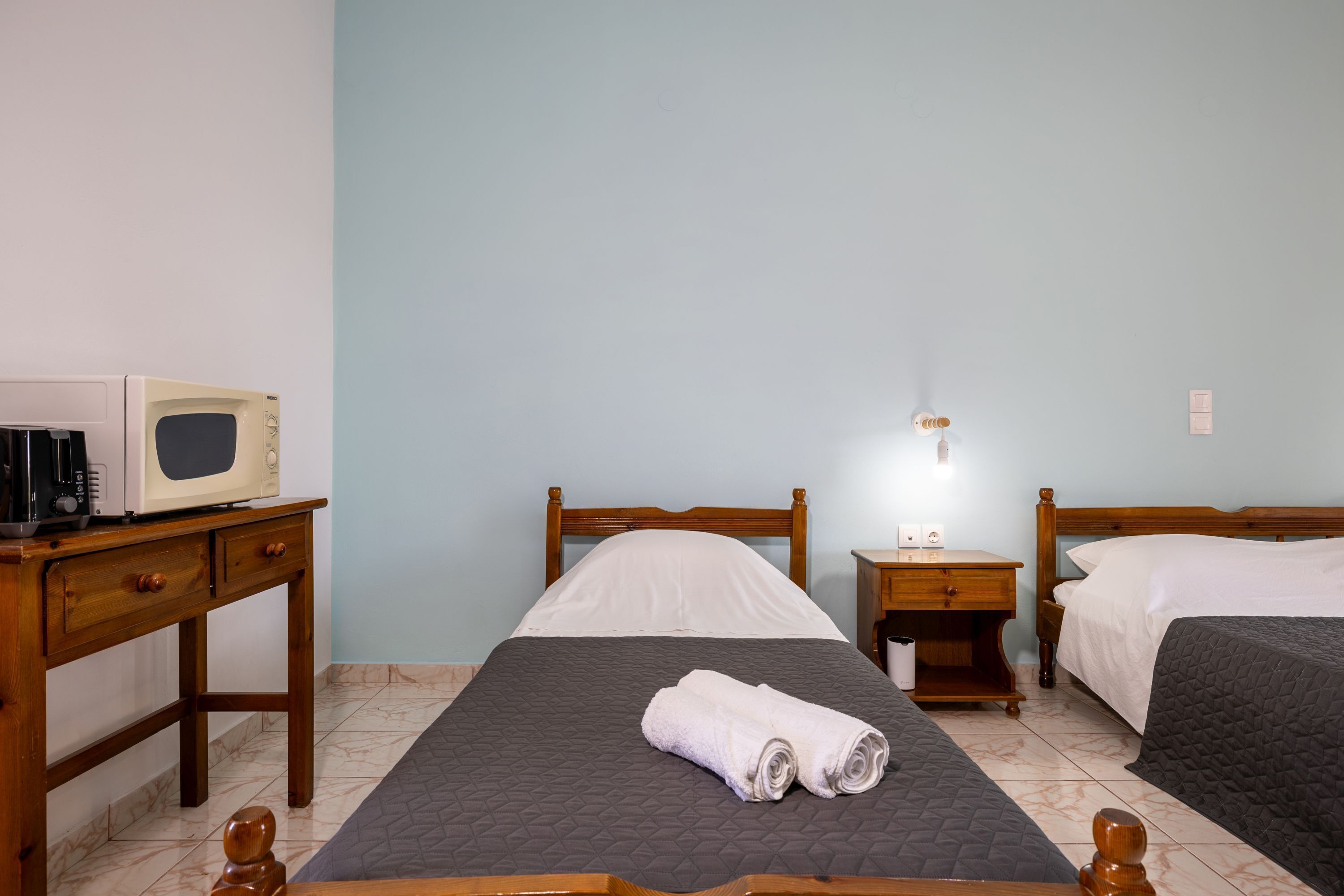 A hotel room with two beds, a wooden desk with a microwave, a nightstand, and a wall-mounted light.