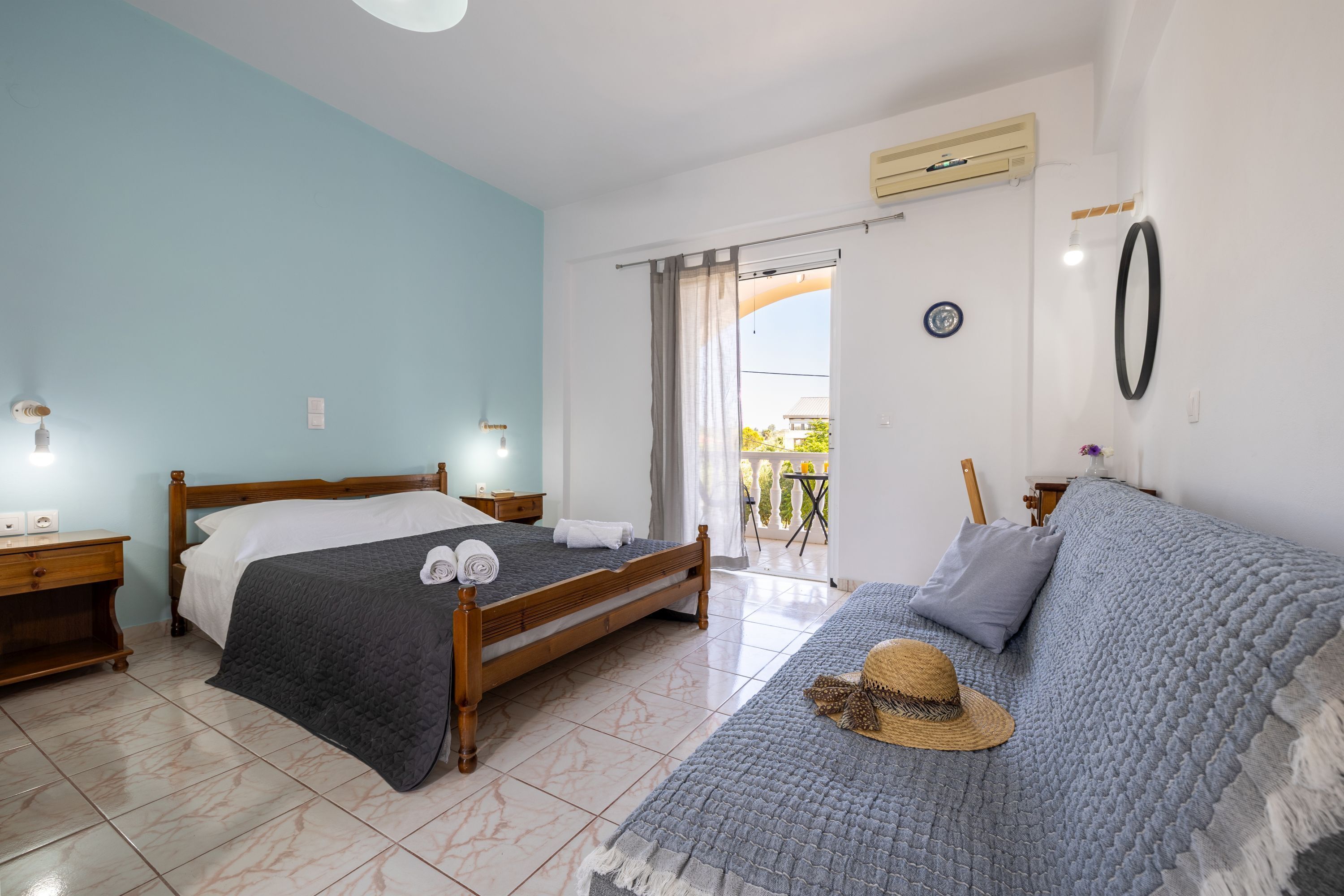 A bedroom with a bed, a sofa, a wooden nightstand, a wall-mounted air conditioner, and a sliding glass door leading to a balcony.