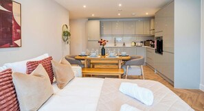 A modern hotel room with a bed, a dining table set for two, and a kitchenette in the background. - FREE Parking, Hi-Speed WiFi,55 Inch 4K TV LongStay (Farnborough)