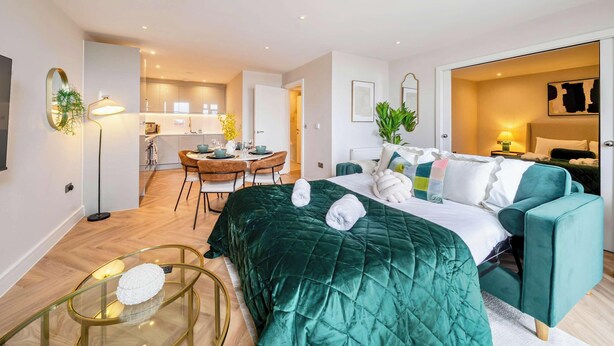 A modern hotel room with a green bed, a sofa, a dining area, and a kitchen in the background.