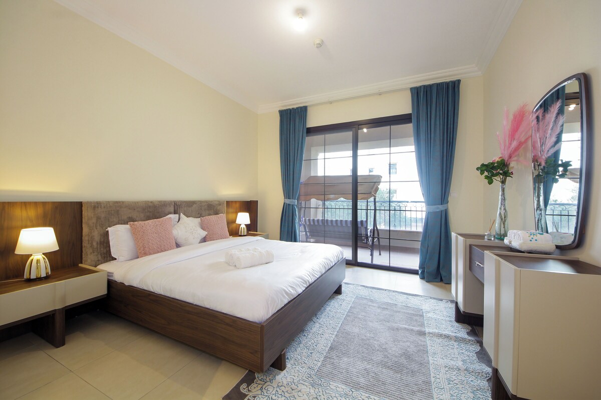 A hotel room with a large bed, a nightstand with a lamp, a dresser, and a balcony with blue curtains.