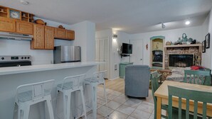 A compact kitchen with a refrigerator, stove, and wooden cabinets. There is a dining area with a table and chairs, and a living room with a fireplace and a television.
