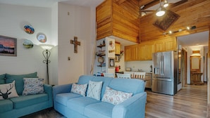 A living room with a blue sofa, wooden flooring, and a kitchen in the background. - Condo within walking distance of the Chile Express (Angel Fire)
