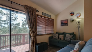 A living room with a sliding glass door to a deck, a green sofa with patterned cushions, and a wall with a 'Friends Gather Here' sign. - Condo within walking distance of the Chile Express (Angel Fire)