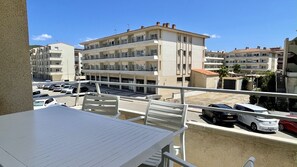 Un balcón con una mesa y sillas, con vista a un estacionamiento con varios coches y un edificio de varios pisos al fondo.