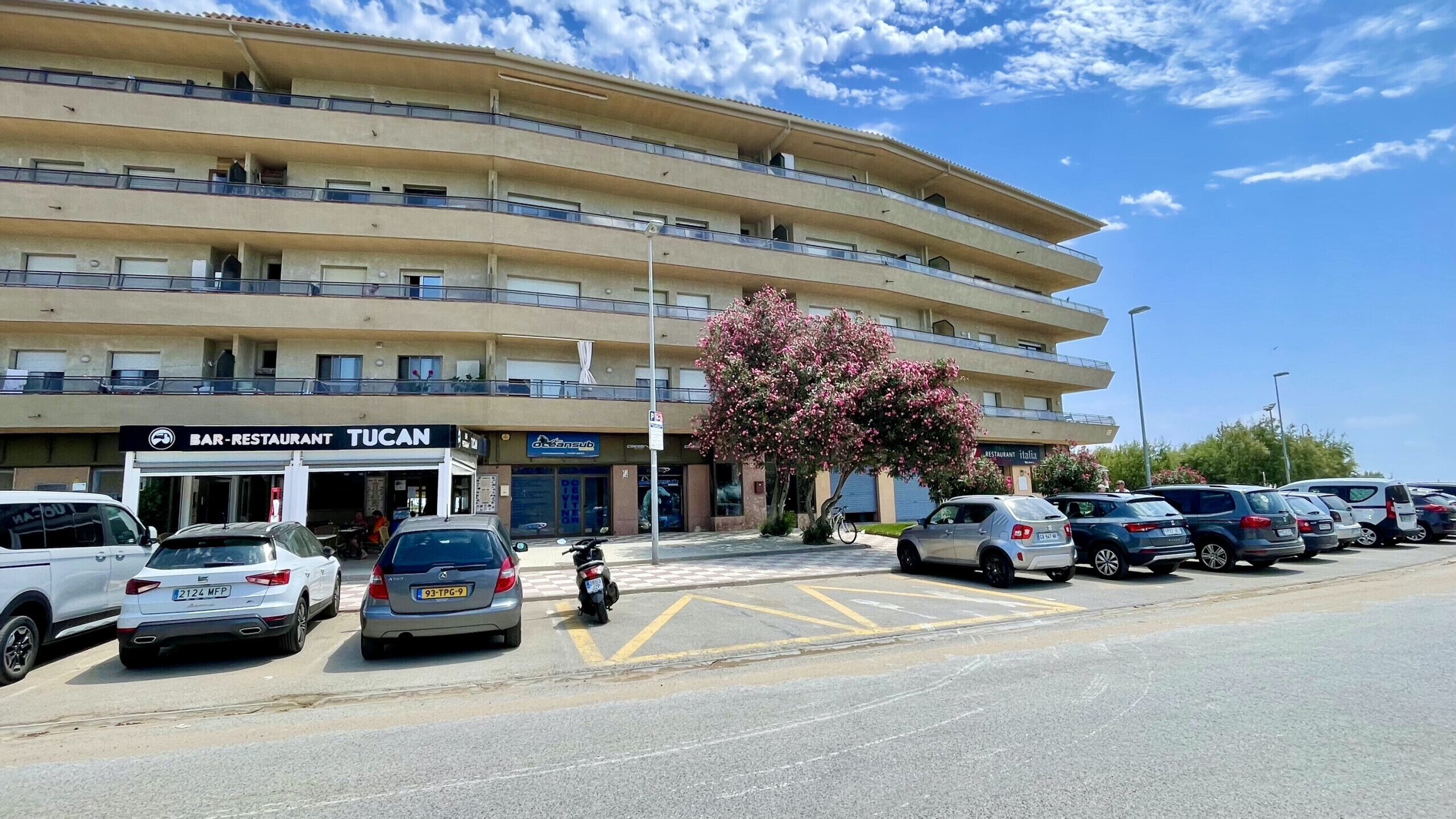 A multi-story building with a bar-restaurant named 'TUCAN' on the ground floor. There are several parked cars and a motorcycle in front.