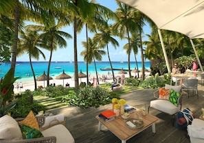 An outdoor seating area with a view of the beach and ocean, featuring palm trees, lounge chairs, and a wooden table with books and drinks.